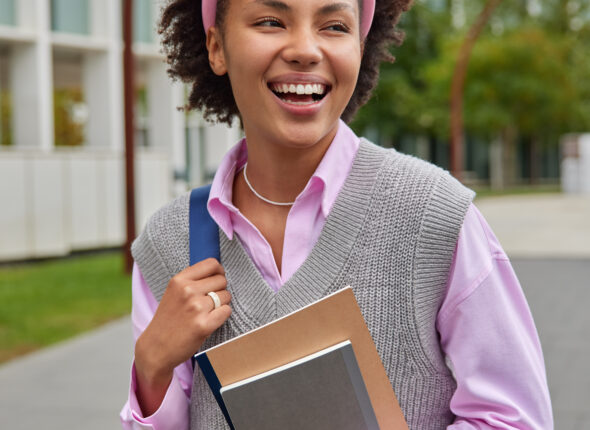 Happy American student strolls in college yard carries rucksack and notepads weas pink shirt knitted vest and hoop surrounded by city street expresses positive emotions. Youth and lifestyle concept