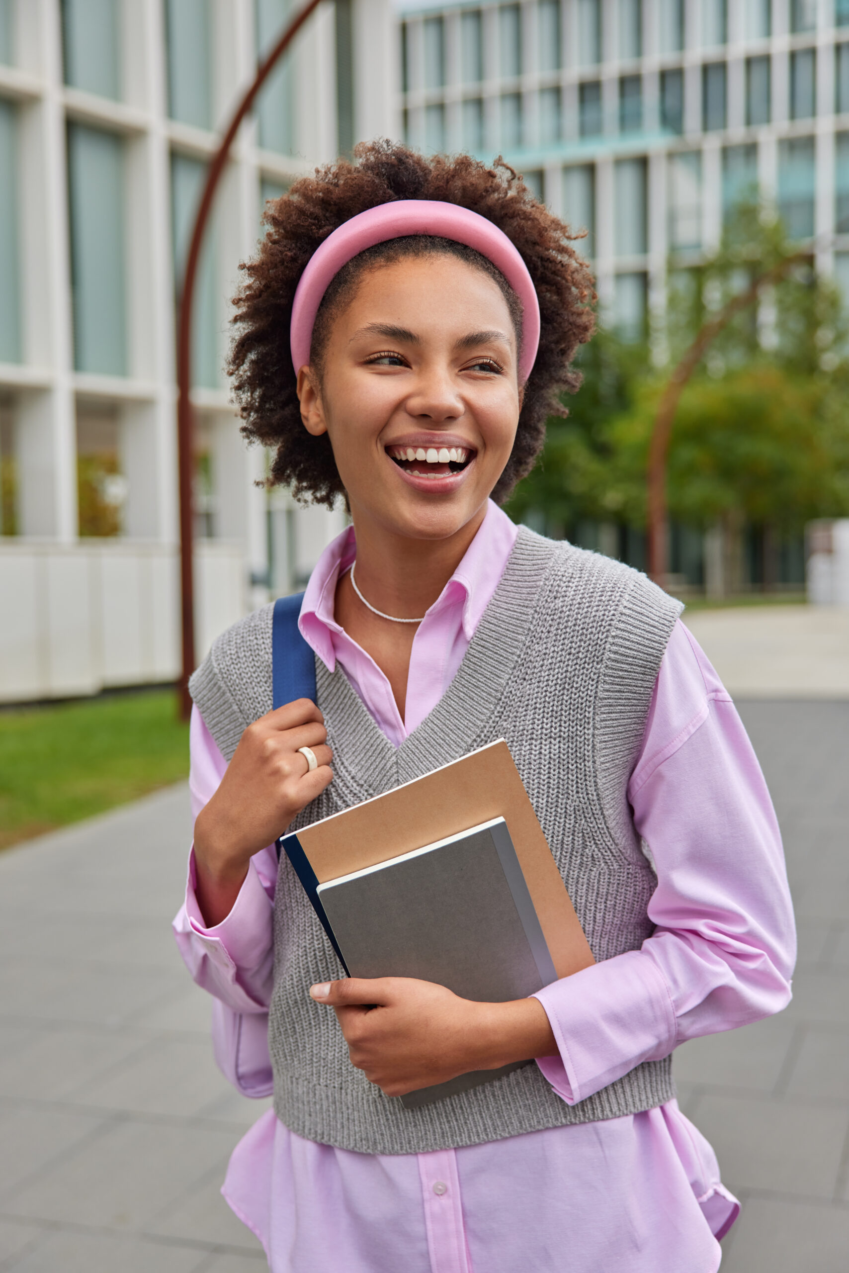 Happy American student strolls in college yard carries rucksack and notepads weas pink shirt knitted vest and hoop surrounded by city street expresses positive emotions. Youth and lifestyle concept